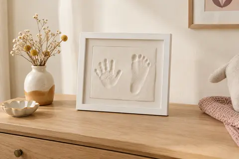 Cadre d’empreinte bébé blanc en bois avec empreintes en argile, posé sur une commode claire.